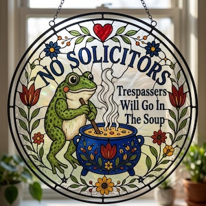 Könnte beinhalten: Ein runder Buntglas-Sonnenfänger mit einem grünen Frosch, der in einem blauen Topf rührt, mit dem Text NO SOLICITORS Trespassers Will Go In The Soup. Das Design umfasst ein rotes Herz, florale Ranken und bunte Blumen in einem schwarzen Rahmen.