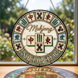 Puede incluir: Una ventana redonda de vidrieras con una cadena dorada para colgar. El diseño presenta la palabra "Mahjong" en cursiva, rodeada de caracteres chinos y las palabras "Social Club". La paleta de colores incluye crema, verde, rojo y dorado.