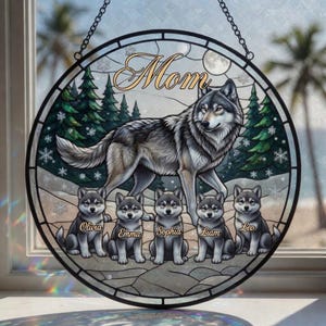 Peut inclure: Vitrail rond représentant un loup gris et cinq louveteaux. Le mot "Mom" est en haut. Les chiots s'appellent Olivia, Emma, Sophia, Liam et Leo. L'arrière-plan comprend des pins et des flocons de neige.