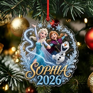 以下が含まれることがあります： 『アナと雪の女王』のエルサ、アナ、オラフが描かれた透明な円形のクリスマスオーナメント。青い雪の結晶、金色の筆記体で書かれた「SOPHIA」という名前、青い文字の「2026」がデザインされています。赤いリボンでクリスマスツリーに吊るされています。