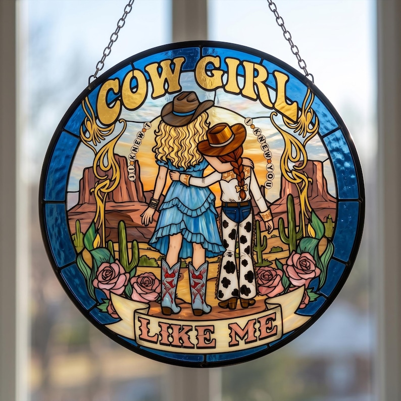 May include: A circular stained glass suncatcher featuring two figures in cowboy hats standing in a desert landscape with cacti and roses. The text reads COW GIRL at the top and LIKE ME at the bottom, with I KNEW YOU written on both sides.