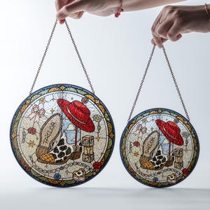 May include: Two circular stained glass suncatchers with hanging chains. The design features a red hat, a cow-print boot with the name Taylor, the number 13, and stacked blocks with the letters T, S, and S. The background includes stars and bows.