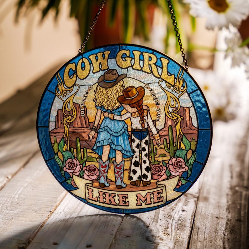 May include: A circular stained glass suncatcher featuring two figures in cowboy hats standing in a desert landscape with cacti and roses. The text reads COW GIRL at the top and LIKE ME at the bottom, with I KNEW YOU written on both sides.
