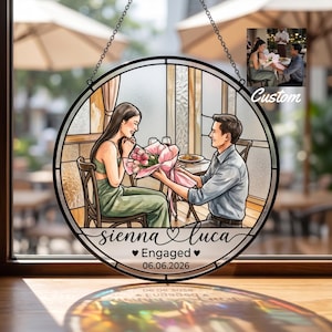 May include: Round stained glass window art featuring a couple at a restaurant, with the man presenting flowers to the woman. The text reads "sienna & luca", "Engaged", and "06.06.2026". The artwork is framed with a black border and hangs from a chain.