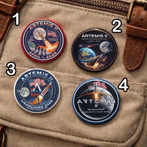 Op de afbeelding: Vier ronde Artemis II missie-spelden. Elke speld toont een raketlancering, de Amerikaanse vlag en tekst. De spelden zijn rood, blauw en wit, met data en zinnen als "Terug naar de Maan" en "Lancering 2026."
