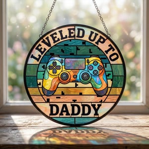 May include: Round stained glass panel with a colorful video game controller design. The panel has the words "LEVELED UP TO DADDY" in black lettering. The controller is in shades of yellow, orange, blue, and green, with a black border and chain for hanging.