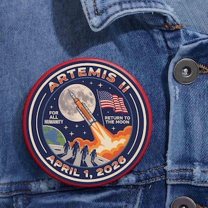May include: A circular patch with a red border featuring the text "ARTEMIS II" and "APRIL 1, 2026." The design includes a rocket launching towards the moon, the American flag, and the words "FOR ALL HUMANITY" and "RETURN TO THE MOON."