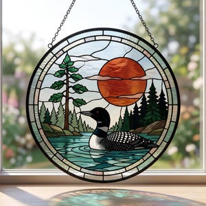 May include: A circular stained glass suncatcher featuring a black and white loon swimming on blue water. The scene includes a tall pine tree, a dense forest of green trees, and a large orange sun in the sky. The piece is framed in black metal.