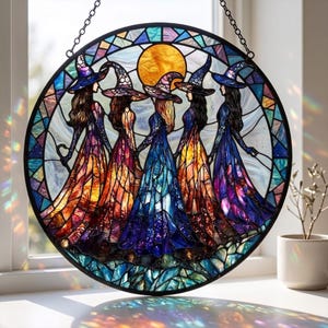 May include: A round stained glass window featuring five witches in colorful dresses and pointed hats, holding hands. The artwork has a vibrant color palette of orange, blue, purple, and yellow, with a sun in the background. The window is framed in black and hangs from a chain.