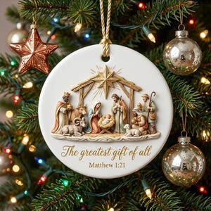 Może przedstawiać: Okrągła biała ceramiczna ozdoba z wypukłą sceną narodzin, złotą gwiazdą, figurkami i zwierzętami. Napis The greatest gift of all oraz Matthew 1:21 wykonano złotą czcionką. Ozdoba zawieszona jest na złotym sznurku.