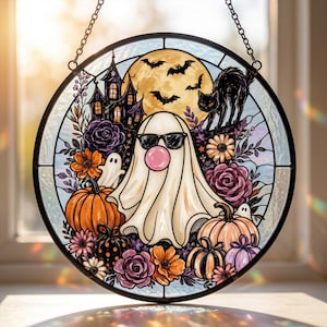 Può includere: Un acchiappasole circolare in vetro colorato raffigurante un fantasma con occhiali da sole e una bolla rosa, circondato da zucche, fiori viola e arancioni, un gatto nero, pipistrelli e la sagoma di una casa stregata in una cornice nera.