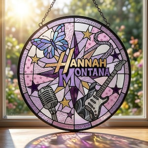 Puede incluir: Vidriera redonda con una mariposa, estrellas, micrófono y guitarra. El texto "Hannah Montana" está en oro y morado. La obra utiliza tonos morados, dorados y blancos, con un borde negro y una cadena.
