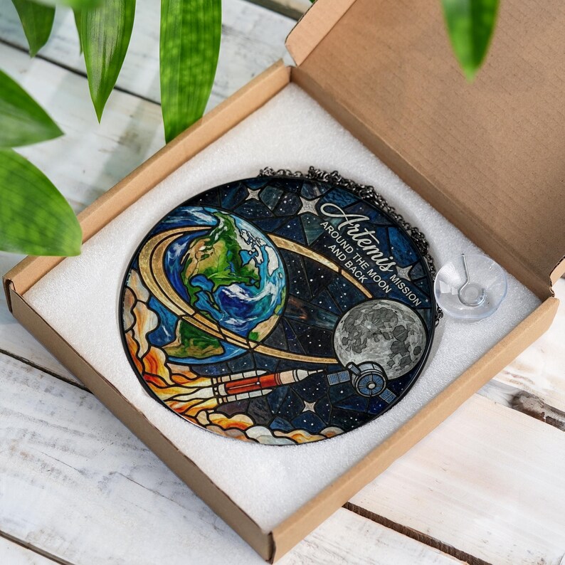 Pu&ograve; includere: Opera d'arte rotonda in vetro colorato raffigurante la Terra, la Luna e un razzo su uno sfondo stellato. Il testo "Artemis Around the Moon and Back" &egrave; visibile. L'opera &egrave; presentata in una scatola di cartone.