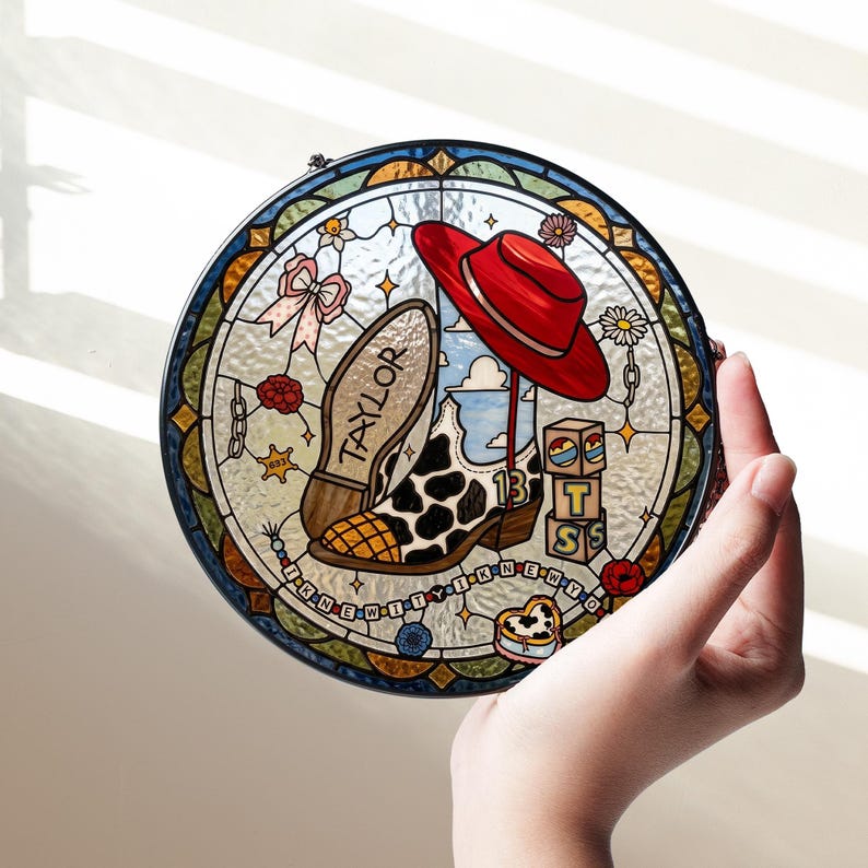 May include: A circular stained glass suncatcher featuring a cow-print cowboy boot with the name TAYLOR, a red hat, and blocks with the letters T, S, and the number 13. The design includes a pink bow, flowers, and the text I KNEW IT Y I KNEW Y O.