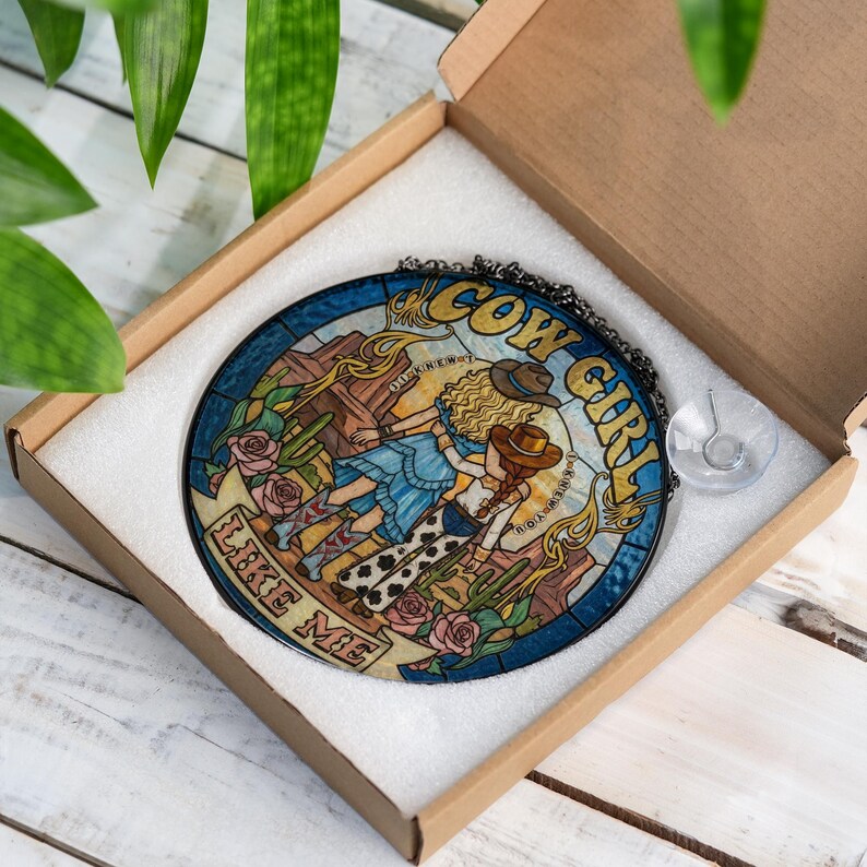 May include: A circular stained glass suncatcher featuring two figures in cowboy hats, desert scenery, and pink roses. The text reads COW GIRL LIKE ME. The piece is framed in black metal with a hanging chain and a clear suction cup, packaged in a box.