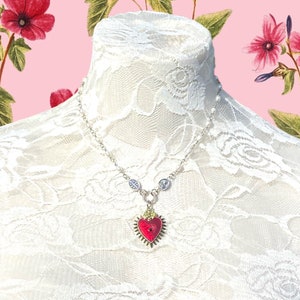May include: A silver chain necklace with a red heart pendant. The heart is detailed with gold accents and a small, clear stone in the center. The necklace is displayed on a white lace background.