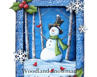 Puede incluir: Una pintura caprichosa de un muñeco de nieve de pie en una escena de bosque nevado. El muñeco de nieve lleva un sombrero de copa negro, una bufanda verde y tiene una sonrisa amigable. El fondo presenta abedules, cardenales rojos y un cielo azul con nieve que cae. La pintura está enmarcada con un borde azul y decorada con copos de nieve y acebo.