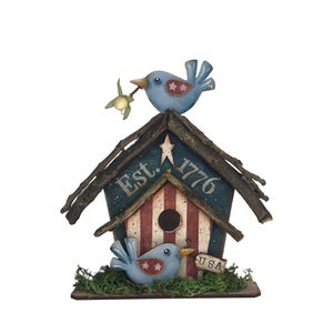 May include: A decorative birdhouse featuring a rustic twig roof, a blue painted front with the text Est. 1776, and red and white vertical stripes. Two light blue birds with star accents are attached, one holding a small flower and one near a USA tag.