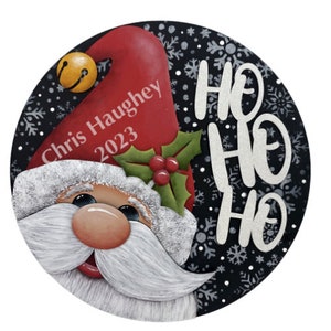 Könnte beinhalten: Ein runder Weihnachtsbaumschmuck mit schwarzem Hintergrund und einem weißen Schneeflockenmuster. Ein Weihnachtsmann-Gesicht mit roter Mütze schaut von der linken Seite des Schmucks hervor. Die Mütze trägt den Namen "Chris Haughey" und das Jahr "2023" darauf geschrieben. Die Worte "Ho Ho Ho" sind in weiß auf der rechten Seite des Schmucks geschrieben.