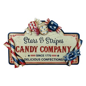 May include: A rectangular metal sign with a cream background featuring the text Stars & Stripes CANDY COMPANY SINCE 1776 DELICIOUS CONFECTIONS. The sign is decorated with three-dimensional metal candy pieces and striped ribbons in red, white, and blue.