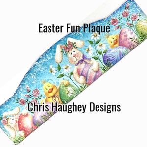以下が含まれることがあります： 青い木製のプレートに、イースターバニーとヒヨコがイースターエッグを持っている絵が描かれています。プレートには「Easter Fun Plaque」と「Chris Haughey Designs」の文字が書かれています。