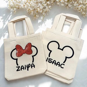 May include: Off-white canvas tote bags with black Mickey Mouse head outlines. One bag features a red glitter bow and the name "ZAIYA". Another bag has the name "ISAAC". The bags have short handles.