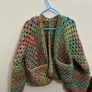Può includere: Giacca all'uncinetto multicolore con due tasche frontali. La giacca presenta un motivo a granny square nei toni del verde, rosa, arancione e marrone. È appesa a una gruccia blu contro un muro bianco.