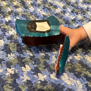 May include: A handmade toy axe with a teal head and a brown handle. The axe head has a black outline around a white rectangle. The axe is held by a person.