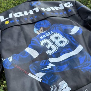 May include: Black leather jacket featuring a hockey player in blue and white, with the name "HAGEL" and the number "38" on the back. The jacket also displays the text "LIGHTNING" and "BE THE THUNDER".