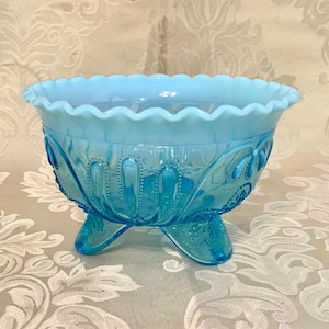 Puede incluir: Un tazón de vidrio azul claro con un borde festoneado y un diseño intrincado. El tazón tiene tres patas y está hecho de vidrio opal.