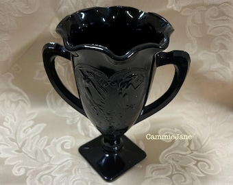 Peut inclure: Un vase en verre noir avec une base carrée et deux poignées incurvées. Le vase a un bord festonné et un motif en relief détaillé. Le vase mesure environ 15 cm de haut.