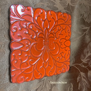 May include: Square, orange ceramic tile with a raised fleur-de-lis design and swirling patterns. The tile has a glossy finish and is likely for decorative use. The tile is a vibrant shade of orange.