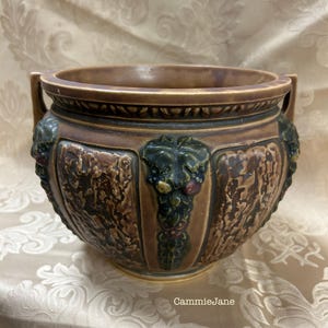 Puede incluir: Una maceta de cerámica marrón con una superficie texturizada y dos asas. La maceta presenta diseños en relieve de vides y caras. El borde y las asas son de un marrón más oscuro, y los detalles en relieve son verdes y marrones.