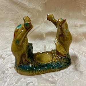 May include: A ceramic frog figurine in shades of green and brown. The figurine has two frogs arching over a small, round dish. The base is a darker green, and the frogs have touches of red and blue. The text "CammieJane" is visible.