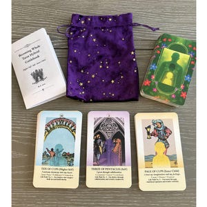 May include: A tarot card set with a guidebook and a purple velvet bag with gold star patterns. The cards feature colorful illustrations and text, including "Ten of Cups," "Three of Pentacles," and "Page of Cups." The guidebook is titled "Becoming Whole Tarot Hybrid Guidebook."
