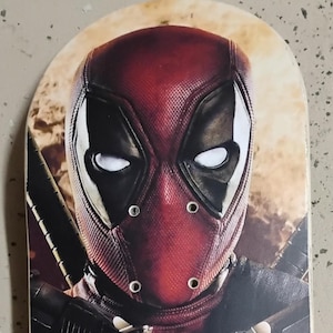 May include: A skateboard featuring a close-up of Deadpool's mask. The mask is predominantly red with black eye cutouts and white eyes. The background has a blurred, fiery effect, and the board has a rounded top.