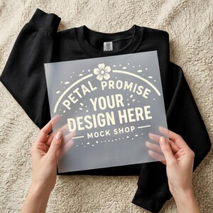 May include: A black crewneck sweatshirt with a transparent sheet overlay. The sheet has the text "Petal Promise Your Design Here Mock Shop" in a light beige color, with a floral design at the top.