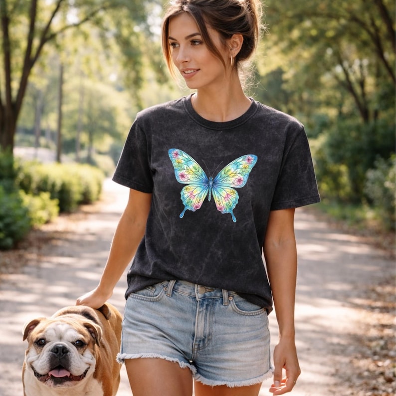 Pu&ograve; includere: Una maglietta grigio scuro con un design a farfalla colorato. La farfalla ha accenti blu, gialli, rosa e verdi. La maglietta &egrave; abbinata a pantaloncini di jeans. Un bulldog cammina accanto.