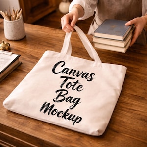May include: A cream-coloured canvas tote bag with the words "Canvas Tote Bag Mockup" in black script. The bag is held by a person, and a stack of books is in the background. The bag is on a wooden surface.