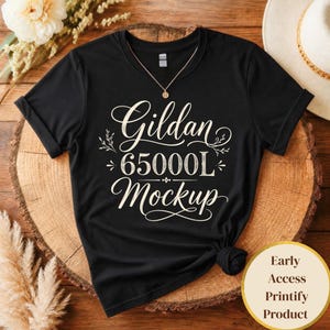 May include: A black Gildan 65000L mockup t-shirt with the text "Gildan 65000L Mockup" in a decorative font. The shirt is displayed on a wooden surface, with a gold necklace and a hat in the background. A round sticker with text is in the lower right corner.
