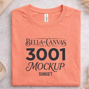 May include: A folded peach-coloured t-shirt with the text "Bella + Canvas 3001 Mockup Sunset" in black script. The shirt is made of a soft material and is a mockup for apparel designs.