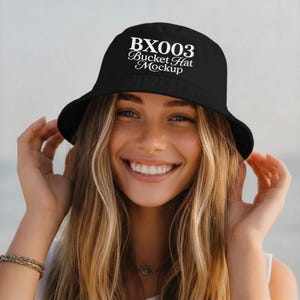 May include: Black bucket hat with the text "BX003 Bucket Hat Mockup" in white. The hat is worn by a person with long blonde hair and a smiling expression. The background is a blurred outdoor scene.