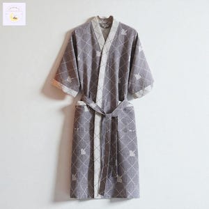 May include: A grey kimono-style robe with a geometric pattern and a matching belt. The robe has white trim along the edges and sleeves. The fabric has a subtle texture, and the design includes small, repeating motifs. The robe is hanging against a white background.