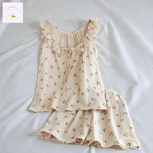May include: A two-piece pajama set in a light cream color featuring a repeating pattern of small dark cherries. The sleeveless top has a ruffled neckline with a tie closure, and the matching shorts have a loose, relaxed fit with a ruffled hem.