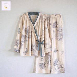 Puede incluir: Un conjunto de pijama beige con una parte superior envolvente y pantalones a juego. El conjunto está adornado con un estampado floral y de mariposas en tonos grises. La parte superior tiene un ribete azul y cierre de lazo.