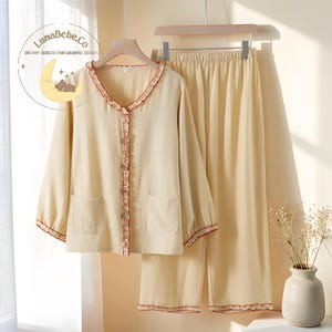 May include: Cream-colored pajama set with red trim. The long-sleeved top has a ruffled collar and button closure, with two front pockets. The matching pants have an elastic waistband. The LunaBebe.Co logo is visible in the upper left corner.