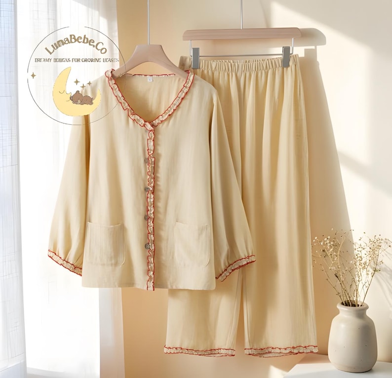 May include: Cream-colored pajama set with red trim. The long-sleeved top has a ruffled collar and button closure, with two front pockets. The matching pants have an elastic waistband. The LunaBebe.Co logo is visible in the upper left corner.