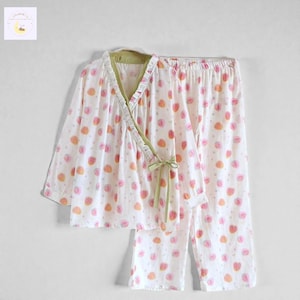 May include: White pyjama set with a strawberry pattern. The top has a wrap-style design with a green trim and a matching tie. The trousers have an elasticated waistband. The strawberries are pink and orange.