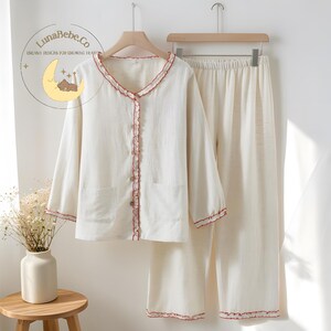 May include: Cream-colored pajama set with red trim. The long-sleeved top has a button-up front, two pockets, and a ruffled edge. The matching pants have an elastic waistband. The LunaBebe.Co logo is visible in the upper left corner.