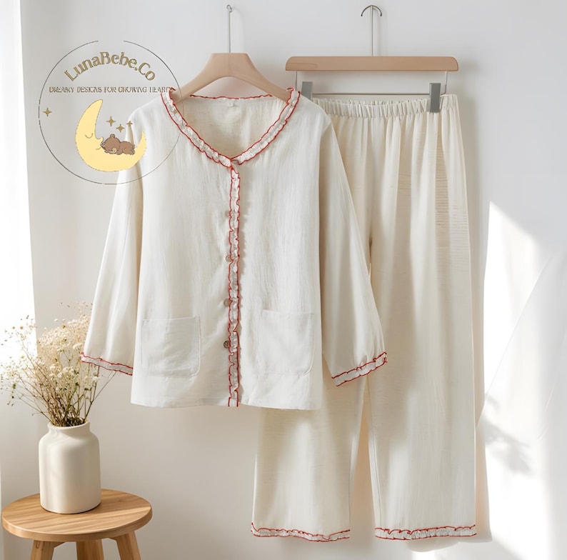 May include: Cream-colored pajama set with red trim. The long-sleeved top has a button-up front, two pockets, and a ruffled edge. The matching pants have an elastic waistband. The LunaBebe.Co logo is visible in the upper left corner.